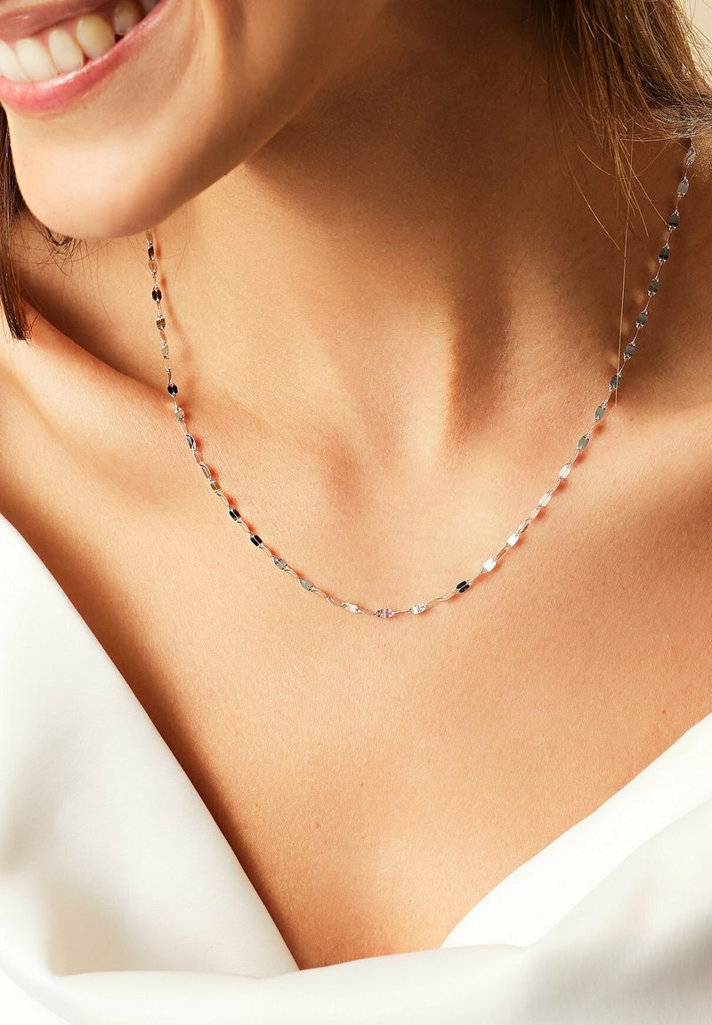 Femme souriante portant un fin collier en argent brillant et un vêtement blanc à épaules dénudées, mettant en valeur son cou et ses clavicules.