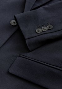 Tissu de costume bleu marine foncé avec trois boutons noirs sur le poignet et un rabat de poche sur le côté, montrant des coutures finement taillées.
