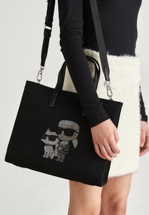 Vrouw draagt zwarte top met lange mouwen en witte gestructureerde rok, houdt zwarte tote tas met cartoonfiguren van strass die een zonnebril dragen.