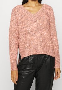 Roze gebreide trui met een V-hals, voorzien van een geribbelde textuur en doorgestikte schouders, gecombineerd met zwarte leren broek.