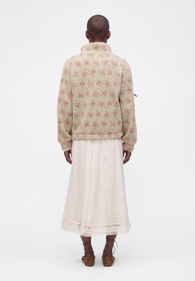 Personne portant une veste légère texturée avec des pois roses, une jupe midi blanche avec des détails en dentelle et des chaussures à lacets marron, vue de dos.