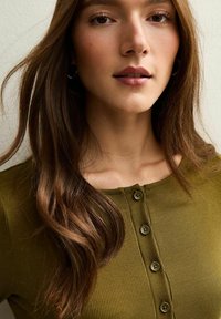 Długorękawowy prążkowany top w oliwkowej zieleni z okrągłym dekoltem i pięcioma guzikami z przodu. Charakteryzuje się dopasowanym krojem i gładką fakturą.