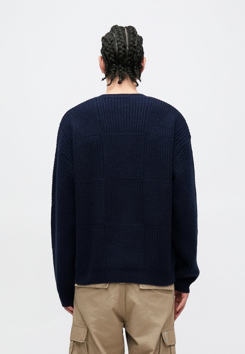 Navy knitted sweater with a textured block pattern, featuring a relaxed fit and wide sleeves, displayed from the back.