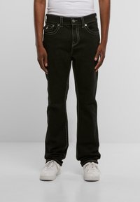 Jeans de mezclilla negra con costuras blancas, diseño de cinco bolsillos, corte recto y cierre de botón. Llevados con zapatillas blancas.