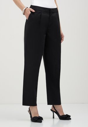 Femme portant un pantalon noir ajusté à longueur de cheville avec des poches latérales, associé à des chaussures à talons hauts noires à bride arrière avec des nœuds.