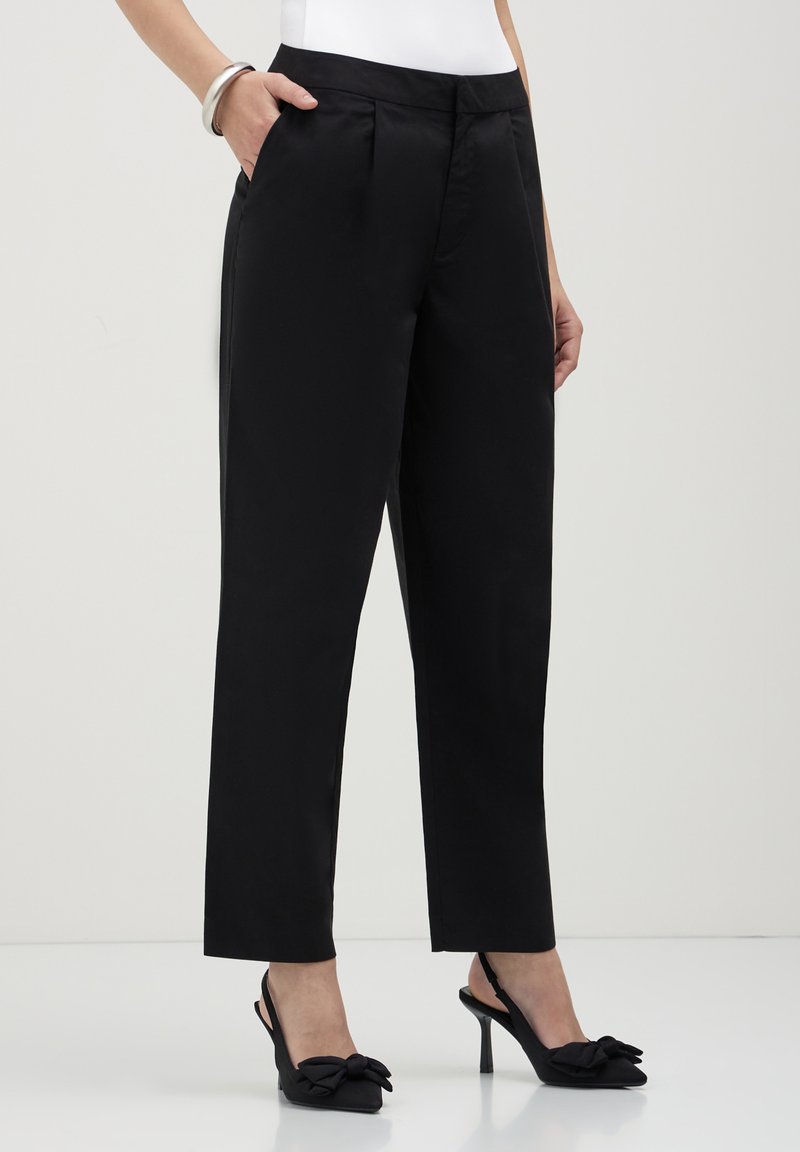 Femme portant un pantalon noir ajusté à longueur de cheville avec des poches latérales, associé à des chaussures à talons hauts noires à bride arrière avec des nœuds.