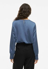 Vrouw met krullend haar, gekleed in een lange mouwen satijnen blauwe blouse met smockmanchetten en zwarte pantalons, met haar rug naar ons gekeerd tegen een effen achtergrond.