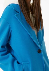 Blå ullblandat överkast med nätt krage, en svart knapp och sidofickor. Slät textur, avslappnad passform och lager med en vit huvtröja.