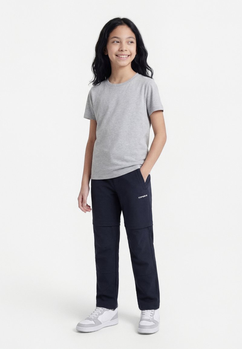 Hosszú fekete hajú lány szürke pólóban, tengerészkék nadrágban és fehér tornacipőben, egyik kezét a zsebébe dugva, mosolyogva, oldalra nézve áll.