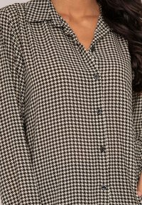 Blouse à motif pied-de-poule noir et blanc avec un col, fermeture à bouton et manches longues. Tissu doux avec une texture lisse.