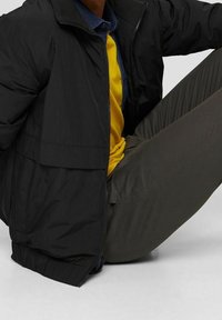 Giacca a vento nera con zip frontale, polsini elasticizzati e superficie liscia. Indossata sopra un maglione giallo e pantaloni grigi.