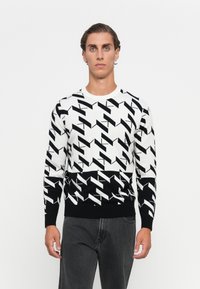 Junger Mann mit kurz zurückgegeltem Haar, der einen schwarz-weißen geometrischen Pullover und dunkle graue Jeans trägt, steht vor einem weißen Hintergrund.
