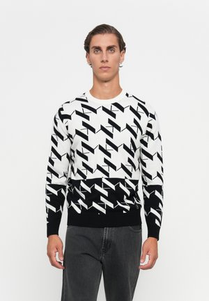 Young man with short slicked-back hair wearing a black-and-white geometric patterned sweater and dark gray jeans standing against a white background.