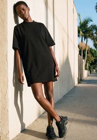 Fiatal személy fekete, túlméretezett pólóruhában és fekete tornacipőben egy napfényes falnak dől egy pálmafákkal szegélyezett járdán.