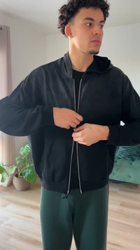 Melnā zip-up džemperis ar kapuci, kam raksturīga smalka tekstūra, elastīgi aproces un priekšējās kabatas, kombinēts ar zaļām biksēm.