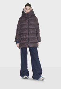 Giubbotto imbottito marrone con colletto alto, chiusura con zip e cuciture orizzontali. Il modello indossa pantaloni larghi in denim scuro e sneakers.