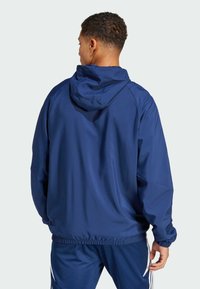 Chaqueta cortavientos de color azul marino con capucha, puños elásticos y dobladillo. Tejido suave, corte holgado y sin logotipos ni patrones visibles.