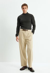 Dunkelgrauer gerippter Rollkragenpullover, beige gefaltete Hose, brauer Gürtel und braune Stiefeletten. Das Model steht vor einem hellen Hintergrund.