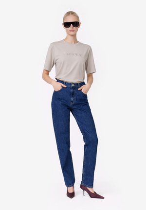 Kobieta ubrana w beżowy t-shirt LAMANIA, niebieskie jeansy z wysokim stanem, ciemne okulary przeciwsłoneczne i bordowe buty na wysokim obcasie, stoi z rękami w kieszeniach.