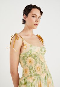 Mouwloze jurk met bloemenpatronen in geel en groen, gerimpelde bovenkant en verstelbare gele strikbandjes. Zachte stoftextuur.