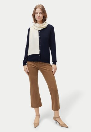 Junge Frau trägt eine marineblaue Strickjacke, weißen Schal, braune verkürzte Hose und beigen Absätzen, steht mit einer Hand in der Hüfte.