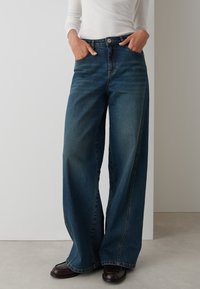 Wide-leg denim jeans in a medium blue wash. Features classic five-pocket design, stitched detailing, and a slightly faded texture.