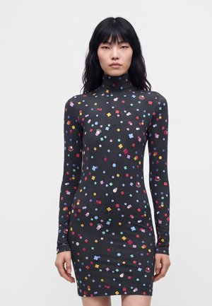 Femme portant une robe noire à manches longues avec col roulé, ornée de formes géométriques colorées dispersées, se tenant devant un fond blanc uni.