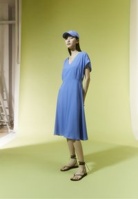 Vestido azul de manga corta y escote en V con una silueta fluida. Combinado con sandalias planas negras con ataduras en los tobillos y una gorra azul a juego.
