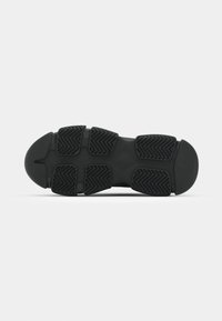 Semelle d'une chaussure de sport noire avec des panneaux de caoutchouc segmentés présentant des motifs de grip en zigzag sur un fond clair.