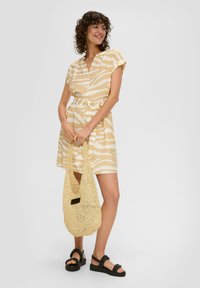 Robe courte à rayures jaunes et blanches avec un décolleté en V, des manches courtes et une taille nouée. Le modèle tient un sac en crochet beige et porte des sandales noires.