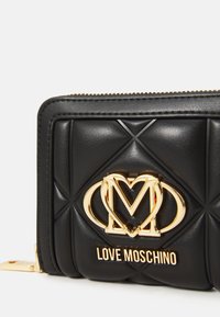 Portofel din piele cu quiltăriță neagră, având un logo auriu în formă de inimă cu litera "M" și textul "Love Moschino", dotat cu un fermoar auriu, pe un fundal alb.