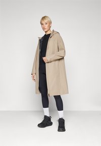 Beige vedenpitävä takki hupulla, jossa on etuvetoketju, rento leikkaus ja sileä pinta, päällä mustat urheiluvaatteet ja kengät.