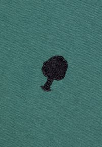 Tessuto di cotone verde con una finitura testurizzata, caratterizzato da un motivo di albero ricamato in nero sul lato sinistro.