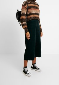 Personne portant un pantalon large noir, un pull en tricot rayé marron et noir, des baskets montantes noires et un sac à dos noir sur fond blanc.