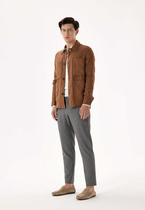 Homme portant une veste marron boutonnée, une chemise blanche, un pantalon gris et des chaussures beiges à enfiler, debout devant un fond clair uni.