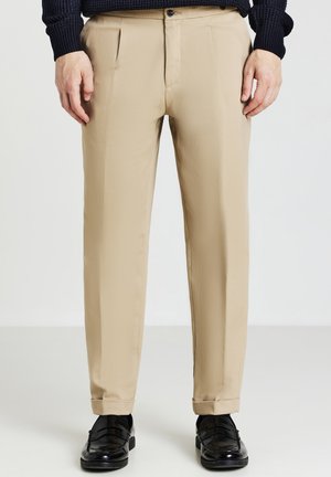 Pantalons beige ajustés avec plis à l'avant, fermeture éclair et bouton unique, associés à des chaussures en cuir noires pour le contraste.