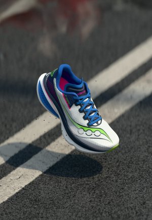 Witte, blauwe en groene Saucony hardloopschoen in de lucht boven een asfaltweg met witte belijning.