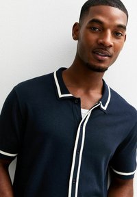 Marineblauw polo-shirt met korte mouwen, voorzien van een witte rand rondom de kraag en de mouwranden. Het shirt heeft een knoopsluiting en een zachte gebreide textuur.
