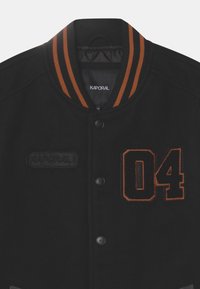 Black varsity jacket with orange striped ribbed collar. Features "Kaporal" logo patch and embroidered number "04." Buttons are black.