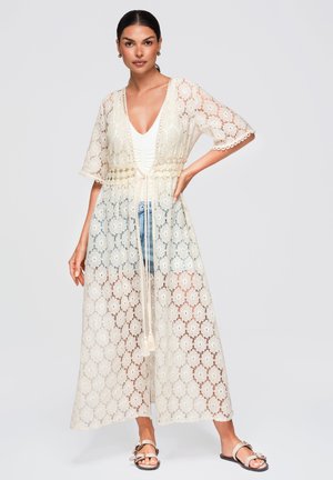Donna che indossa un lungo kimono in pizzo crema con motivi floreali sopra una canottiera bianca e shorts in denim, abbinato a sandali beige a ciabatta.