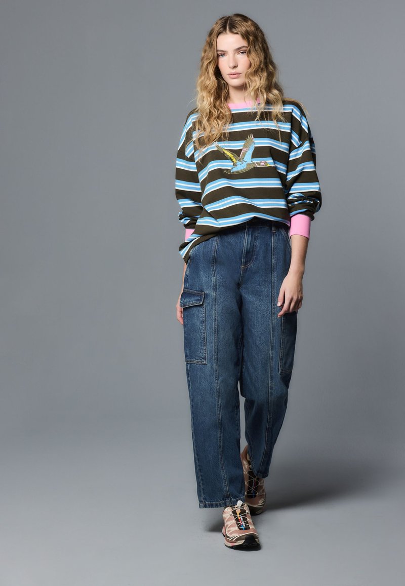 Vrouw met golvend blond haar staat, draagt een gestreepte trui met vogelmotief, wijde denim cargobroek en sneakers met roze accenten op een grijze achtergrond.