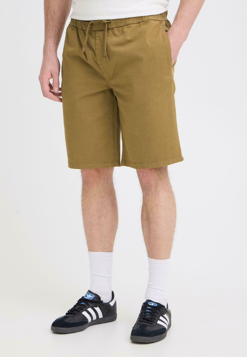 Homme portant un short à cordon de couleur moutarde, des chaussettes blanches montantes et des baskets Adidas noires avec des rayures blanches sur un fond blanc.