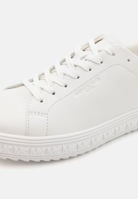 Baskets basses blanches avec lacets, tige en cuir lisse, logo embossé sur le côté, et bord de semelle à motif avec initiales répétées.