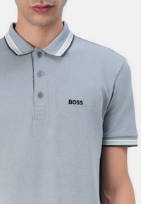 Világosszürke polo póló, textúrált anyagból, gombos kivágással és fekete "BOSS" logóval. A gallér fehér és sötétszürke csíkokkal díszített.