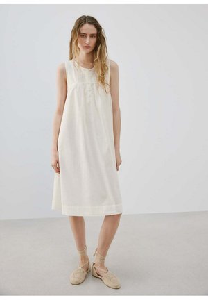 Kobieta ubrana w białą, bezrękawnikową sukienkę za kolano z guzikami i beżowe szydełkowe espadryle, stojąca na jednolitym tle podłogi i ściany.