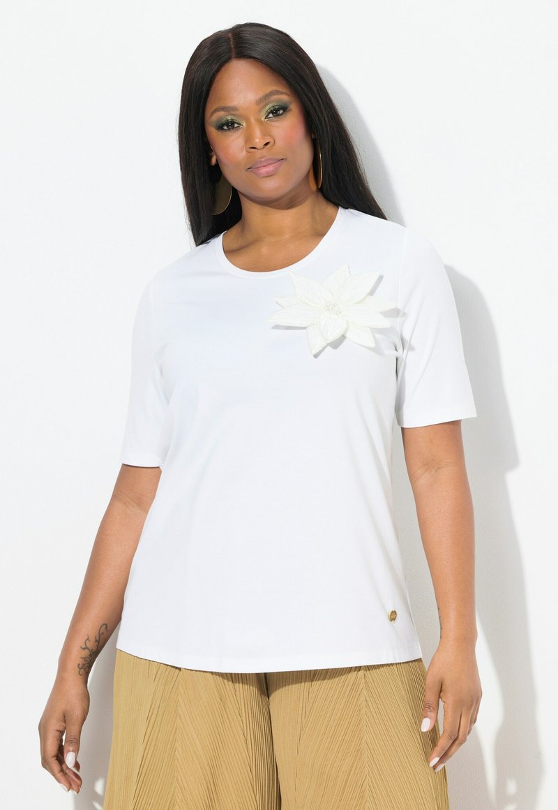 Top blanco de manga corta con cuello redondo y un detalle floral texturizado blanco en el pecho. Combinado con pantalones plisados de color beige.