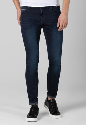 Mann trägt dunkelblaue Slim-Fit-Jeans, die an den Knöcheln umgekrempelt sind, ein weißes T-Shirt und schwarze Sneakers mit weißen Sohlen und steht auf einem grauen Hintergrund.