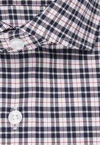 Colletto e parte anteriore della camicia bianca con motivo a scacchi nero e rosso e bottoni bianchi.