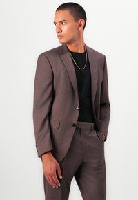 Abito marrone a scacchi con un design aderente, chiusura a un bottone e due tasche, abbinato a una maglietta nera a girocollo e una collana d'oro.