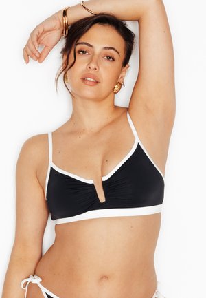 Donna che indossa un top bikini nero con bordi bianchi, alza il braccio sinistro, accessoriata con orecchini a cerchio d'oro e braccialetti, su sfondo bianco.